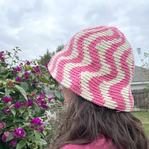 Puede incluir: Un sombrero de cubo de crochet a rayas rosa y blanco. El sombrero lo lleva una persona con el pelo largo castaño y una camisa rosa con estampado.
