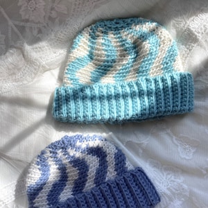May include: Two crocheted hats with a white and blue chevron pattern. The hat on the top is light blue and the hat on the bottom is a darker blue.