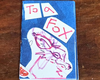 May include: A small, handmade card with a blue background and the words "To a Fox" in red. The card features a whimsical drawing of a fox in pink and white, with simple line art. The overall design is playful and artistic.
