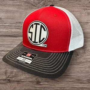 May include: Red and white trucker hat with a black brim and white mesh back. The hat features a white circular patch with the letters "STC" in black. The brim has white stitching and a "Richardson" tag.