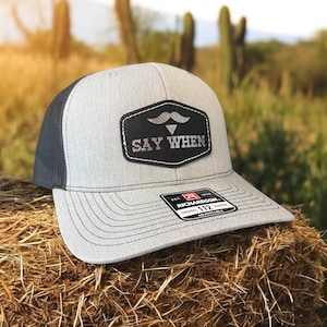 May include: A gray and black trucker hat with a black leather patch that says "SAY WHEN" with a silver mustache above the text. The hat is sitting on a bale of hay.