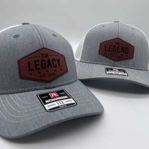 May include: Two gray and white trucker hats with brown leather patches. The patch on the left hat says "The Legacy Est. 2018". The patch on the right hat says "The Legend Est. 1954".