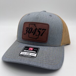 May include: Gray trucker hat with a brown leather patch featuring the Georgia state outline and the number "30457". The hat has a mesh back and a curved brim. The Richardson brand tag is visible.