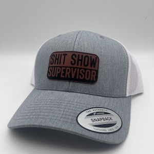 May include: Gray and white trucker hat with a brown leather patch that says "Shit Show Supervisor". The hat has a YUPOONG logo and the text "The Authentic Snapback Yupoong Since 1974".