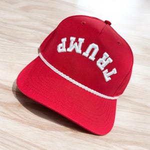 May include: Red baseball cap with a white rope trim and white embroidered text that reads "TRUMP" in an arched design.