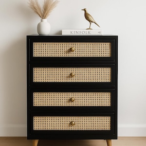 May include: A black four-drawer chest with woven cane drawer fronts and brass knobs. The chest is topped with a white vase, dried grass, a gold bird figurine, and a book titled "KINFOLK". The chest has light wood legs.