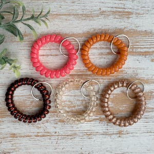 May include: Five spiral keychain rings in various colors: pink, orange, brown, clear, and gold. Each ring has a silver-colored key ring attached. The keychains are arranged on a light-colored wooden surface.