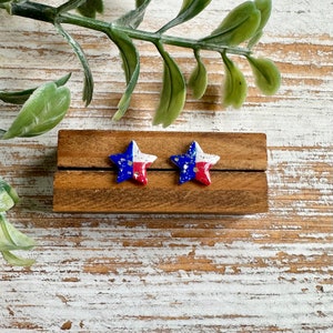 May include: A pair of star-shaped earrings with a white, red, and blue paint design. The stars are displayed on a wooden surface.