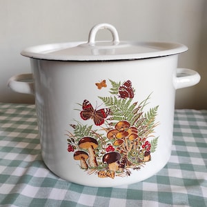 Op de afbeelding: Witte emaille pot met een deksel en zijhandvatten. De pot heeft een kleurrijke illustratie van paddenstoelen, varens en vlinders. De pot staat op een groen-wit geruit tafelkleed.