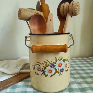 Pode incluir: Um recipiente de esmalte cor de creme com borda preta, uma alça de arame com empunhadura de madeira e um decalque floral de margaridas brancas e flores azuis. O recipiente está cheio de vários utensílios de cozinha de madeira, incluindo colheres.