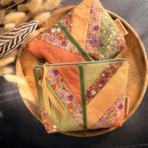 Könnte beinhalten: Zwei kleine, gesteppte Stoffbeutel mit einem Patchwork-Design in Orange-, Grün- und floralen Mustern. Die Beutel haben einen Reißverschluss und ein kleines Stoffetikett. Sie werden auf einem Holztablett präsentiert.
