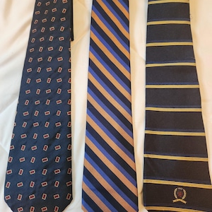 May include: Three neckties displayed side by side. The left tie is dark blue with small red-outlined rectangular patterns. The center tie features diagonal stripes in blue and gold. The right tie is dark blue with horizontal gold stripes and a crest.