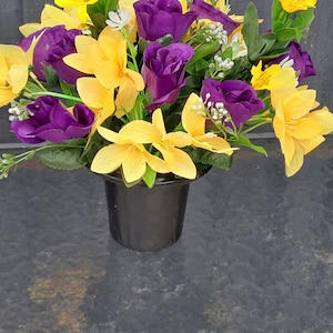 May include: A bouquet of yellow and purple artificial flowers in a black pot. The flowers are arranged in a cluster and are in full bloom.