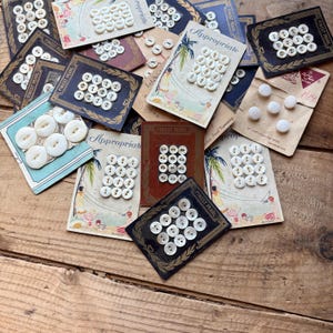 May include: A collection of vintage button cards featuring various white, round, four-hole buttons attached to illustrated cardboard backing. The cards display the text Appropriate, Finest Pearl, and Le Chic, showcasing assorted button sizes and styles.