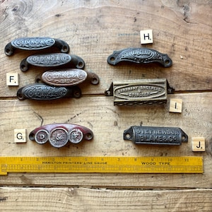 May include: A collection of metal drawer pulls and bin handles with embossed text including Olding & Co, D&C Co, Barnhart Brothers & Spindler, and Stephenson Blake & Co. A yellow Hamilton Printers' Line Gauge ruler is positioned at the bottom.