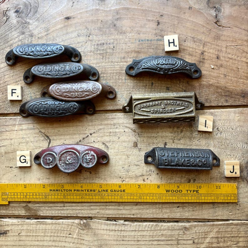 May include: A collection of metal drawer pulls and bin handles with embossed text including Olding & Co, D&C Co, Barnhart Brothers & Spindler, and Stephenson Blake & Co. A yellow Hamilton Printers' Line Gauge ruler is positioned at the bottom.