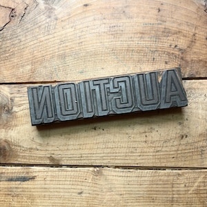 May include: A rectangular metal printing block featuring the word AUCTION in raised, bold, sans-serif capital letters. The metal surface has a dark, weathered finish with visible horizontal line textures within each letter, mounted on a wooden base.