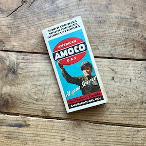 Puede incluir: Un mapa vintage de American Oil Company para Carolina del Norte, Carolina del Sur, Georgia y Florida. El mapa presenta un diseño azul y blanco con el logotipo de Amoco Gas y un hombre con uniforme sosteniendo una boquilla de bomba de gasolina. El texto dice "American Amoco Gas At Your Service from Maine to Florida American Oil Co."