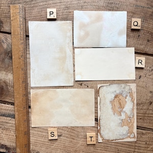 May include: A collection of vintage ephemera, including aged paper rectangles and a wooden ruler, arranged on a wooden surface. The papers show signs of age, with stains and discoloration. Scrabble tiles with letters and numbers are scattered around the papers.