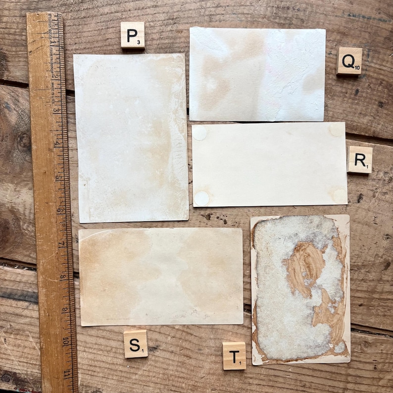 May include: A collection of vintage ephemera, including aged paper rectangles and a wooden ruler, arranged on a wooden surface. The papers show signs of age, with stains and discoloration. Scrabble tiles with letters and numbers are scattered around the papers.