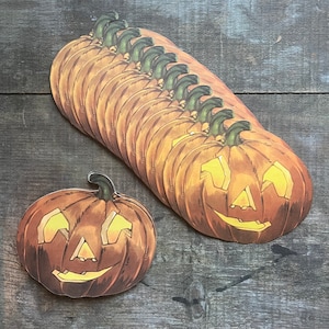 May include: A stack of 10 orange paper jack-o'-lantern cutouts with black eyes, noses, and mouths. The jack-o'-lanterns are arranged in a semi-circle with the largest jack-o'-lantern at the bottom.
