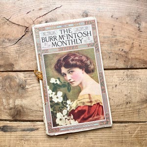 May include: Vintage "The Burr McIntosh Monthly" magazine from May 1909. The cover features a portrait of a woman with flowers, framed by a decorative border. The magazine is on a wooden surface.