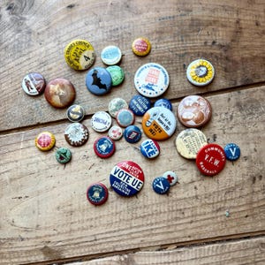 Könnte beinhalten: Eine Sammlung von Vintage-Ansteckknöpfen mit verschiedenen Designs und Texten. Die Knöpfe zeigen Bilder einer Fledermaus, einer Sonnenblume und Texte wie "VOTE UE" und "I LIKE IKE". Die Knöpfe sind auf einer Holzoberfläche verstreut.