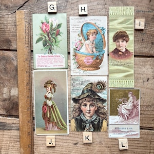 May include: A collection of vintage advertising cards and ephemera, including a card with a floral illustration and text, a card with a child in a basket, and a card with a woman's portrait. The items are arranged on a wooden surface.