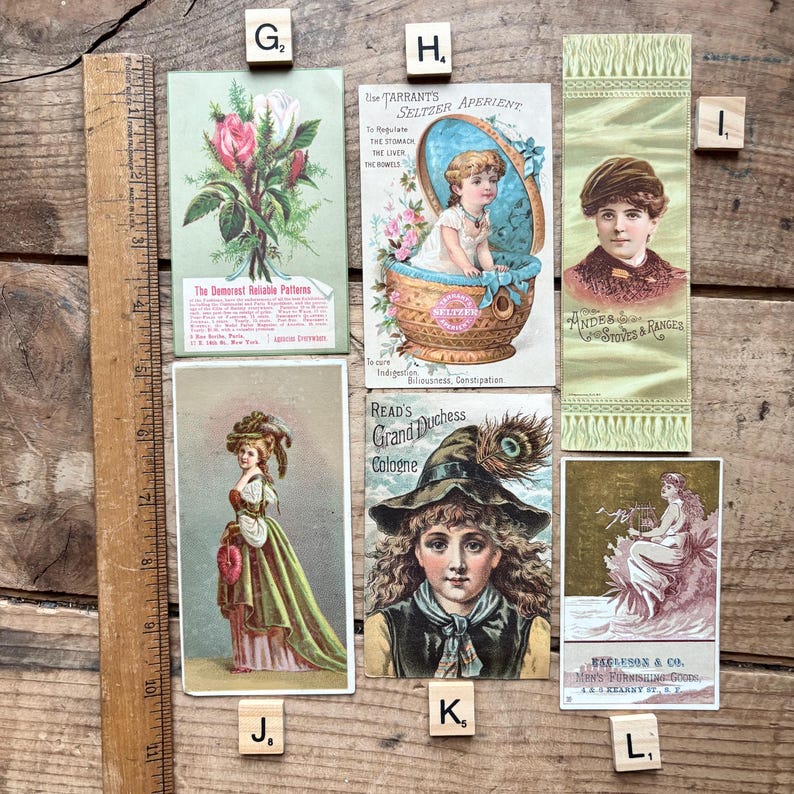 May include: A collection of vintage advertising cards and ephemera, including a card with a floral illustration and text, a card with a child in a basket, and a card with a woman's portrait. The items are arranged on a wooden surface.