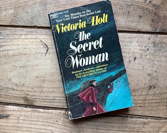 Puede incluir: Un libro de bolsillo titulado The Secret Woman de Victoria Holt con una portada azul oscuro y turquesa. La portada muestra a una persona con capa roja cerca de la proa de un barco. El texto incluye Six Months on the New York Times Best Seller List.