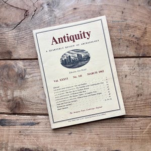 Peut inclure: Un exemplaire vintage de "Antiquity", une revue trimestrielle d'archéologie, datant de mars 1962. La couverture présente le titre en bordeaux, un dessin d'une structure en pierre et les détails de la publication. Le magazine est sur une surface en bois.