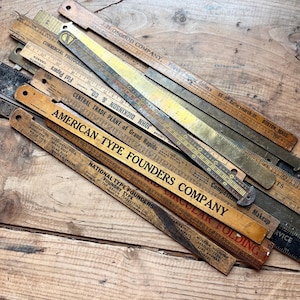 Könnte beinhalten: Eine Sammlung alter Lineale aus Holz und Metall sowie Setzschiffe für Drucker. Die Gegenstände tragen aufgedruckte Texte wie J.H. Osgood Company, American Type Founders Company und National Type Founders sowie verschiedene Maßangaben.