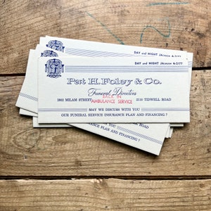 以下が含まれることがあります： 青と白の文字が書かれた、ヴィンテージの名刺の束。「Pat H. Foley & Co. Funeral Directors」と連絡先情報が記載されています。風化した木製の表面に置かれています。
