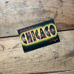 May include: Vintage postcard with the words "Greetings from CHICAGO ...at Night" in a stylized font. The word "CHICAGO" is in yellow with images of the city at night. The postcard has a black background and is on a wooden surface.