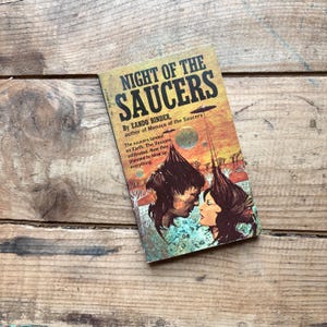 May include: Vintage paperback book titled "Night of the Saucers" by Eando Binder. The cover features a retro sci-fi illustration of a couple and flying saucers. The book is set against a wooden background.
