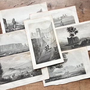 May include: A selection of antique black and white landscape prints. The prints feature scenes of figures in mountainous areas, buildings, and natural landscapes. The prints are displayed on a wooden surface, highlighting their aged paper and detailed illustrations.