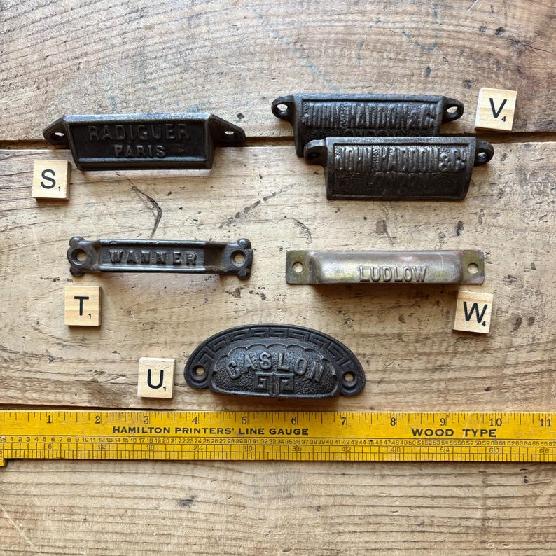 May include: A collection of five metal drawer pulls labeled Radiguer Paris, John Haddon & Co, Wanner, Ludlow, and Caslon, arranged with small lettered tiles S, T, U, V, and W, positioned above a yellow Hamilton Printers' Line Gauge ruler.