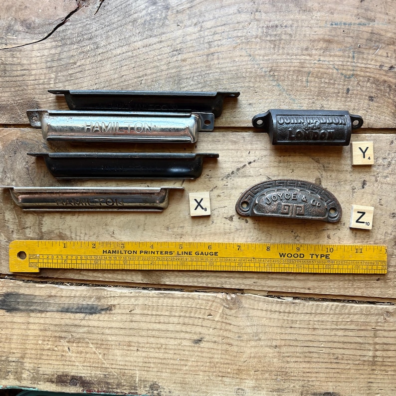 May include: A collection of six metal drawer pulls and handles of varying shapes and finishes, including two labeled Hamilton, one John Haddon London, and one Joyce & Co. A yellow 11-inch Hamilton Printers Line Gauge ruler is placed below the items.