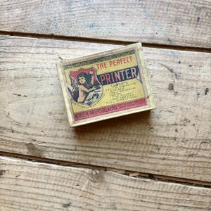 Puede incluir: Una caja de cartón rectangular para el set The Perfect Printer, con una ilustración vintage de una figura alada. La etiqueta incluye el texto: The Perfect Printer, Contents: 4 A Font Rubber Type, 1 Set Rubber, 1 Shell Indelible Ink, 1 Ink Pad, 1 Type Holder, 1 Pair Tweezers, Full Directions, Robt. H. Ingersoll & Bro. 65 & 67 Cortlandt St. New York City.