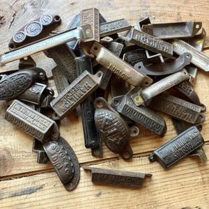 Pode incluir: Uma coleção de puxadores de gaveta e alças de metal vintage em vários acabamentos, incluindo ferro fundido, latão e cromo. Muitas peças apresentam texto em relevo como Hamilton, Ludlow e The Tubbs Mfg Co, algumas com padrões decorativos.