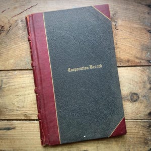 Könnte beinhalten: Ein Vintage-Buch "Corporation Record" mit dunkelgrauem Einband und weinrotem Rücken und Ecken. Der Titel ist in goldfarbenen Buchstaben gehalten. Das Buch liegt auf einer verwitterten Holzoberfläche.