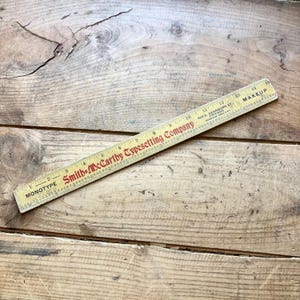 May include: A vintage wooden ruler measuring 15 inches in length. The ruler features black markings and red text that reads Smith-McCarthy Typesetting Company, Monotype, and 508 S. Dearborn St. Chicago. It includes a scale for makeup measurements.