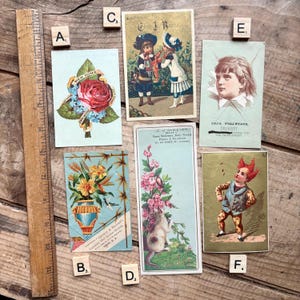 May include: A collection of antique postcards, featuring floral designs, children, and illustrations. The cards showcase a variety of styles, including a rose with a banner, a vase of flowers, and a child's portrait. The cards are arranged on a wooden surface.