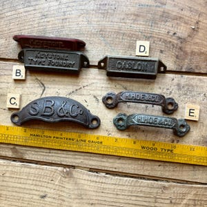 May include: A collection of five vintage metal drawer pulls with embossed text including Keystone Type Foundry, Caslon, SB&Co, and R. Hoe & Co. A yellow Hamilton Printers' Line Gauge ruler is positioned below, showing measurements in inches.