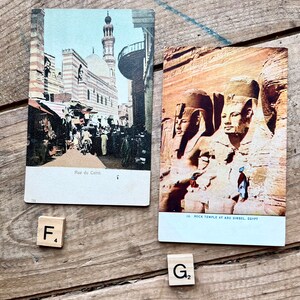 May include: Two vintage postcards depicting scenes from Egypt. The first postcard shows a street scene in Cairo with a mosque in the background. The second postcard shows the rock temple at Abu Simbel with the four colossal statues of Ramses II.