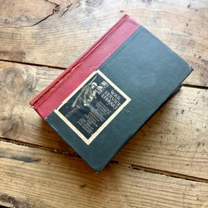 Può includere: Un libro antico della War Service Library. Il libro ha il dorso rosso e la copertina verde scuro. La copertina presenta un'immagine in bianco e nero di un soldato e il testo "War Service Library".