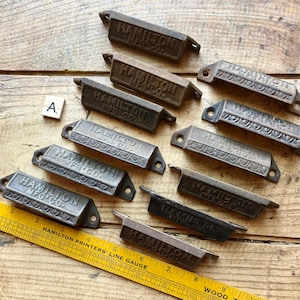 May include: A collection of ten dark metal drawer pulls, each embossed with the text HAMILTON MFG CO. The pulls feature a rectangular design with mounting holes on the ends. A yellow wooden ruler is positioned at the bottom for scale.