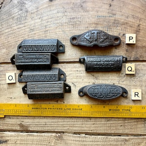 May include: A collection of six dark metal drawer pulls with various embossed text and patterns, including The Tubbs Mfg Co, Anglo Canadian, and M & W Mfg Co, arranged near a yellow Hamilton Printers' Line Gauge ruler measuring up to 11 inches.