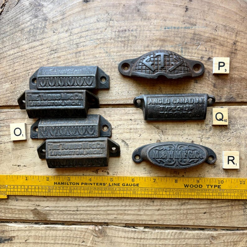 May include: A collection of six dark metal drawer pulls with various embossed text and patterns, including The Tubbs Mfg Co, Anglo Canadian, and M & W Mfg Co, arranged near a yellow Hamilton Printers' Line Gauge ruler measuring up to 11 inches.