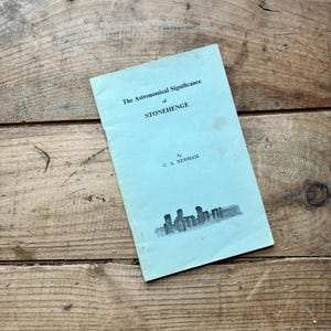 May include: A vintage book titled "The Astronomical Significance of Stonehenge" by C. A. Newham. The book has a light blue cover with black text and a line drawing of Stonehenge. The book is on a wooden surface.